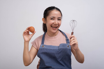A smiling woman in an apron holds an egg in one hand and a whisk in the other, ready to cook or bake.