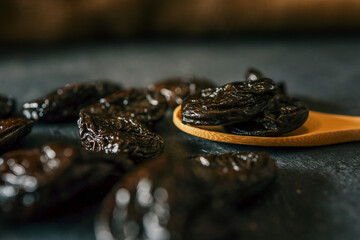 Close-up of glossy dried prunes placed on a dark textured surface, with a few prunes resting on a...
