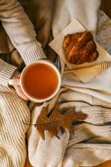 Warm autumn scene with hands holding cup of tea, croissant on parchment, honey dipper, dried oak leaf, and cozy knitted blankets in soft beige tones
