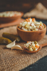 Close-up of traditional Olivier salad with peas, ham, potatoes, carrots, and eggs served in a rustic wooden bowl and spoon on burlap cloth with garlic and pickles on a dark background