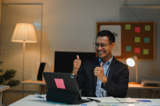 Asian businessman showing thumbs up during video call on tablet in office at night
