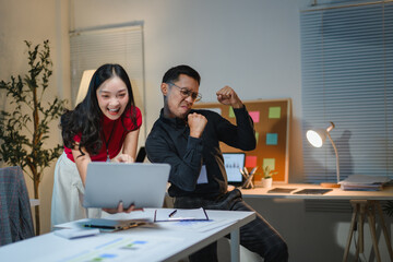 Asian business team celebrating success, looking at laptop in office at night