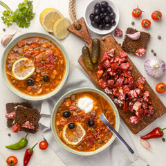 Traditional solyanka soup with smoked sausage, lemon slices, black olives, pickles, and rye bread, top view on white rustic table, cozy daylight