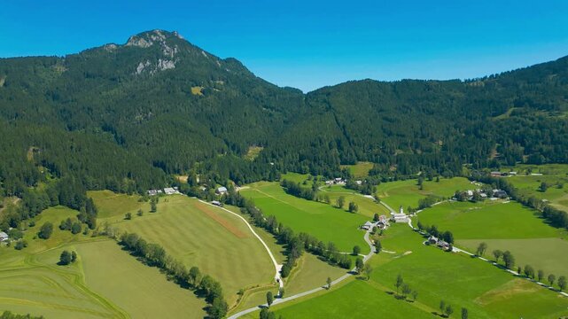 Aerial panoramic view of Zgornje Jezersko valley with lush green meadows, winding roads, and surrounding Kamnik-Savinja Alps under a clear summer sky in Slovenia. Scenic mountain landscape