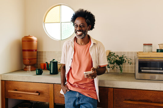 Mature man smiling and standing in a Brazilian kitchen, using smartphone