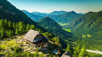 Aerial view of the Češka koča mountain lodge at Spodnje Ravni, surrounded by lush green mountains and forests of the Kamnik-Savinja Alps in Slovenia. Popular destination for hiking, trekking