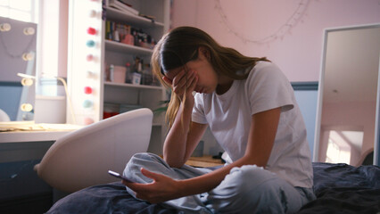 Depressed Teenage Girl Sitting On Bed At Home Looking At Mobile Phone With Head In Hands
