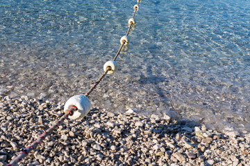 Beach safety concept. A buoy marker line on a calm pebble beach with transparent water on a sunny summer day.Swimming area boundary line with floats on a beautiful pebble beach on the Adriatic coast