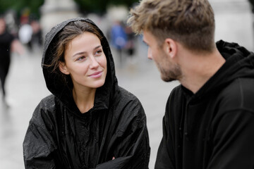 Fototapeta premium A young couple is seen engaged in a warm discussion while sheltering from the rain under hoods, showcasing connection and intimacy in an urban setting.