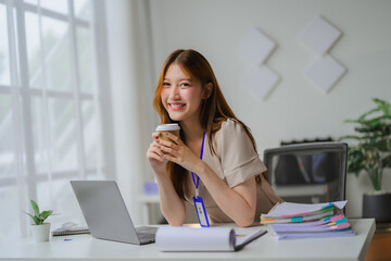 Obraz premium Young businesswoman smiling and holding coffee cup while working on laptop in modern office