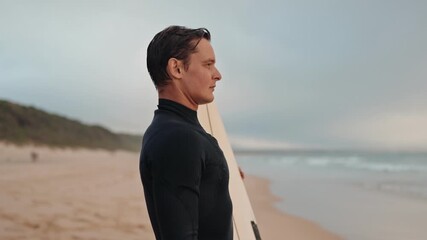 A surfer in a black wetsuit stands on a beach holding his surfboard and staring thoughtfully at the ocean waves.