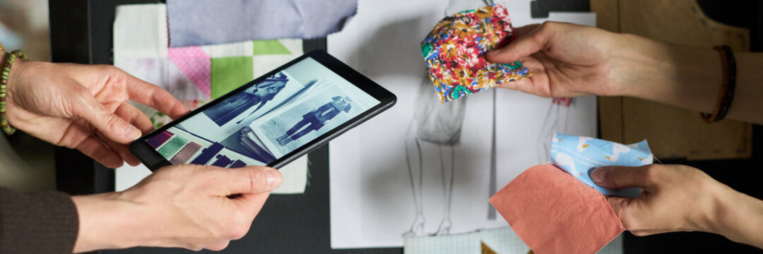 Caucasian woman holding tablet displaying fashion sketches while another Caucasian woman selecting colorful fabric swatches, collaborating on clothing design project at workspace - Powered by Adobe