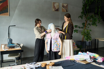 Two Caucasian young adult women collaborating while dressing mannequin with patchwork clothing in...