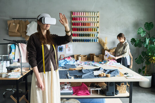 Young adult Caucasian woman wearing virtual reality headset standing in fashion design studio gesturing with hand, while another young adult woman working with fabric in background