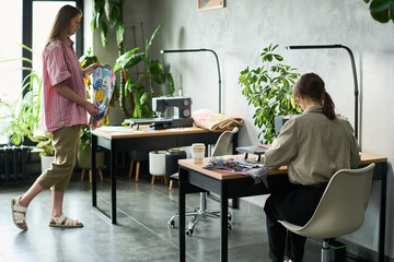 Caucasian young adult woman standing holding colorful fabric, while another Caucasian young adult woman sitting working at sewing machine in modern workspace with plants visible