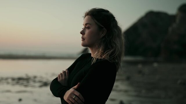 A thoughtful woman stands with arms crossed, gazing into the distance at sunset on a beach. The serene atmosphere evokes contemplation and introspection.