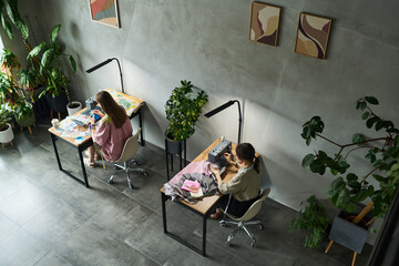 Two young adult Caucasian women sitting at separate desks working on creative projects, one drawing with colored pencils, other using sewing machine, surrounded by indoor plants