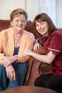 A visiting care worker and an elderly woman sit smiling on a sofa comfortable in each others company with big smiles for the camera.