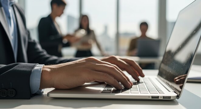 Focused professional typing on laptop in modern office with colleagues collaborating in background