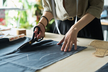 Caucasian young adult woman cutting fabric with scissors on worktable, using pattern pieces for garment making, measuring tape hanging around neck, hands focusing on precise tailoring