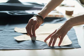 Caucasian young adult man marking fabric with tailor chalk while holding paper pattern, hands visible working on textile, preparing material for garment construction in workshop