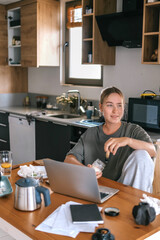 Young woman working remotely at home kitchen table with laptop, coffee, and breakfast, representing...