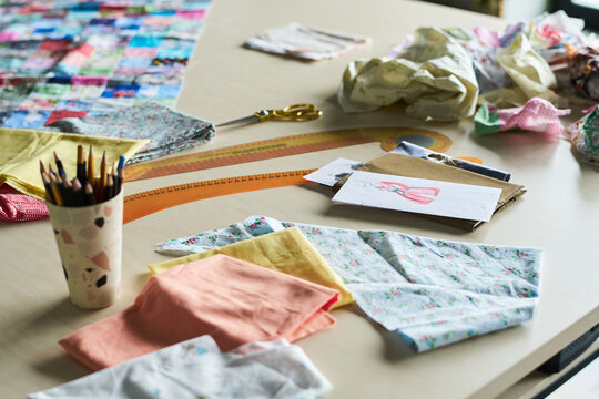 Assorted fabric swatches, colored pencils, scissors, measuring tape, and fashion sketch scattered on table suggesting textile design process or garment creation in progress