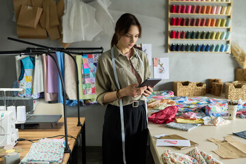 Young adult Caucasian woman standing in sewing studio holding digital tablet, measuring tape around neck, surrounded by colorful fabrics and sewing supplies, focusing on screen