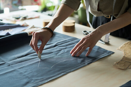 Caucasian young adult woman marking fabric with tailors chalk on worktable, hands drawing pattern lines for garment making, sewing tools and paper pattern pieces visible in workspace
