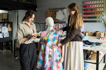 Caucasian young adult woman holding clipboard observing Caucasian woman adjusting colorful patchwork fabric on dress form in modern sewing studio with thread spools visible