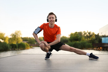 Smiling sportsman in headphones and armband doing morning exercise on pier near river