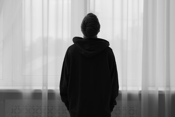 Silhouette of sad man near window at home, back view. Black and white effect