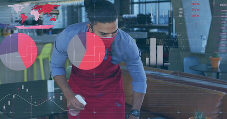 Cleaning server wearing red apron and mask spraying table with bottle at café, with charts