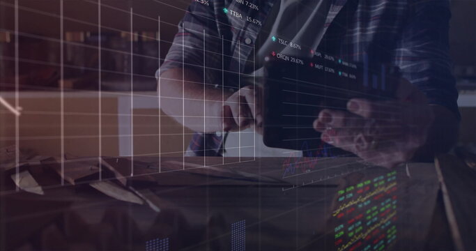 Interacting middle-aged man in checkered shirt tapping tablet on desk in office, stock data overlay