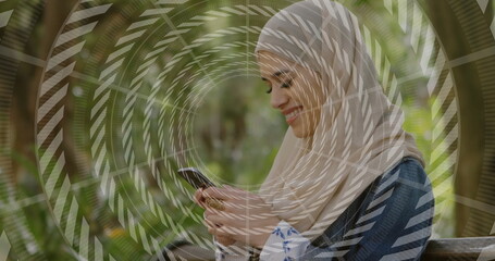 Smiling woman wearing denim jacket and headscarf reading smartphone on bench in park, with rings