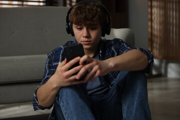 Teen problems. Upset teenage boy in headphones with smartphone near sofa at home