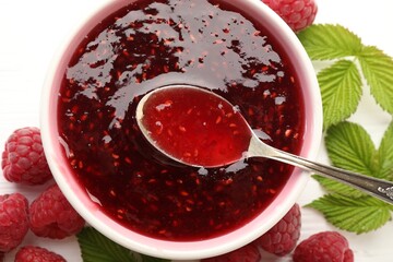 Sweet raspberry jam in bowl, berries and green leaves on white table, flat lay