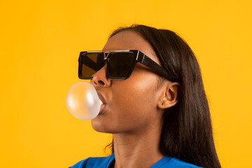 Model blowing bubblegum bubble while wearing sunglasses on yellow background