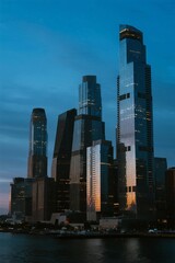 Obraz premium A stunning view of modern skyscrapers illuminated at blue hour along an urban riverfront, showcasing architectural grandeur and city lights.