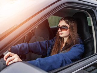 A woman is driving a car with her sunglasses on. She is wearing a blue hoodie and a pair of...
