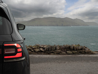 Black car parked with view on amazing nature scene with ocean, mountain, and cloudy sky. Dingle peninsula, Ireland. Popular travel area with stunning Irish landscape. Nobody.