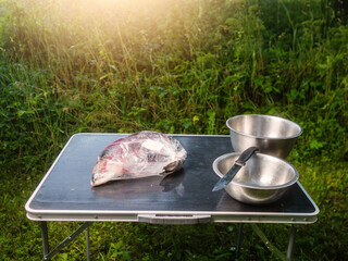 Fresh salmon in a plastic bag on a camping table with two stainless steel bowls for bones and meat. Cooking fine fish product. Preparation stage. Green grass background. Sun flare.