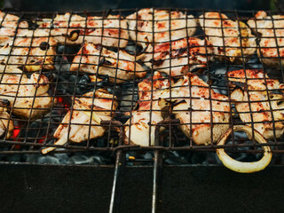 A grill full of meat is cooking over a fire. The meat is being cooked on a metal grate, and the smoke from the fire is rising up. The scene is lively and energetic. Summer time party food preparation