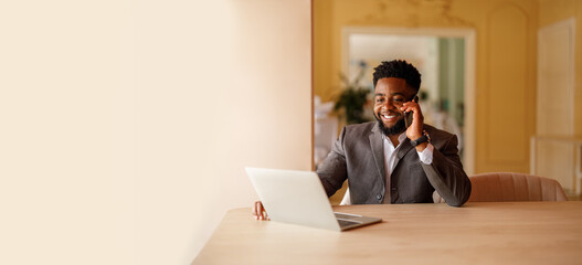 Smiling young businessman discussing over mobile phone while using laptop at desk by wall in modern office