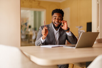 Cheerful male manager with note pad and laptop on desk discussing strategies over mobile phone in...