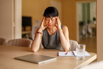 Exhausted professional woman with head in hands and eyes closed sitting at corporate desk in office