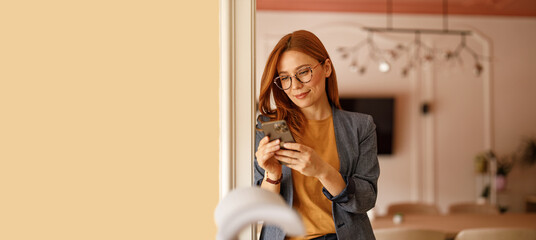 Confident businesswoman in eyeglasses messaging over smart phone while standing by wall in modern...