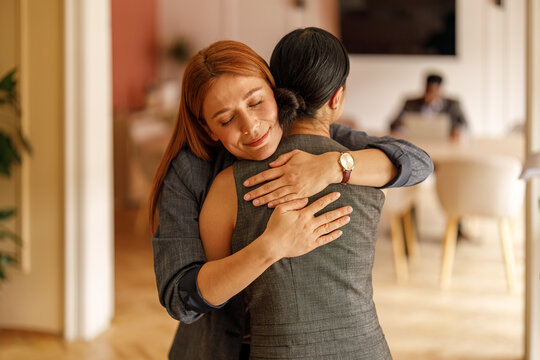 Businesswoman with eyes closed embracing female coworker offering comfort and motivation in corporate workplace