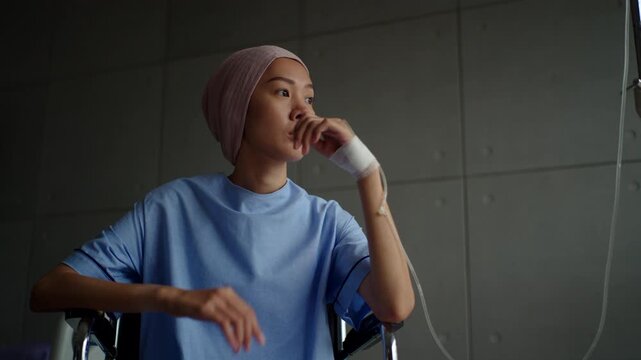 Depressed patient woman with cancer sitting on wheelchair suffering headache
and looking out the window after chemotherapy at hospital, sickness female wearing head scarf feeling unwell and lonely.