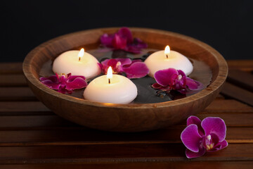 Burning candles and orchid flowers in bowl of water on wooden table against black background,...